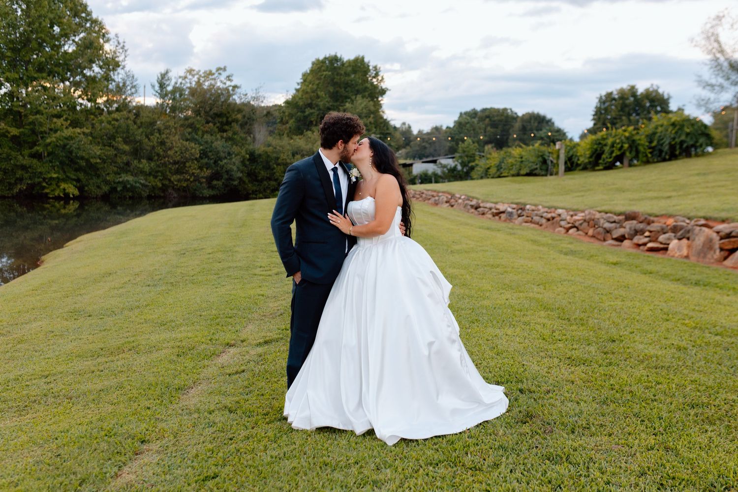 Bride and groom share a romantic kiss on a lush green lawn near a pond on their wedding day.