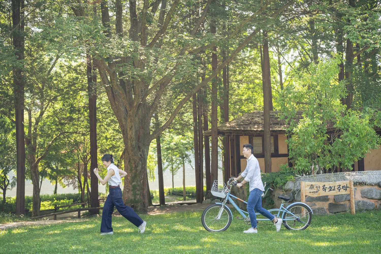 Two people walking and cycling through a lush green park surrounded by tall trees near a small wooden pavilion.