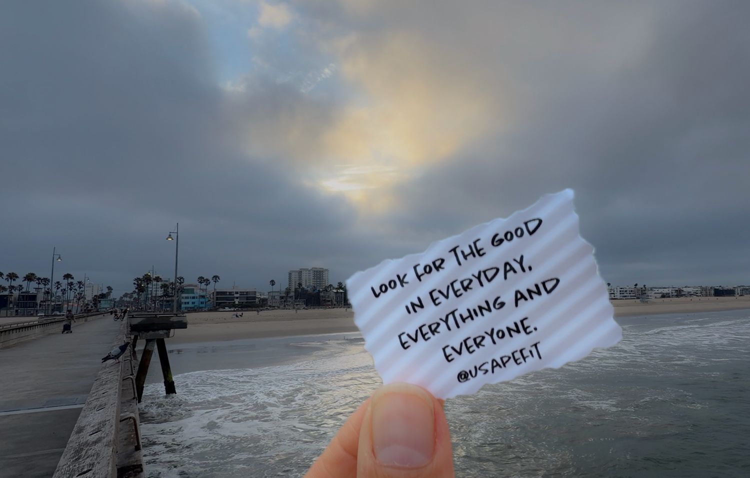 Hand holding a note reading 'Look for the good in everyday, everything and everyone' on a beach pier at sunset.