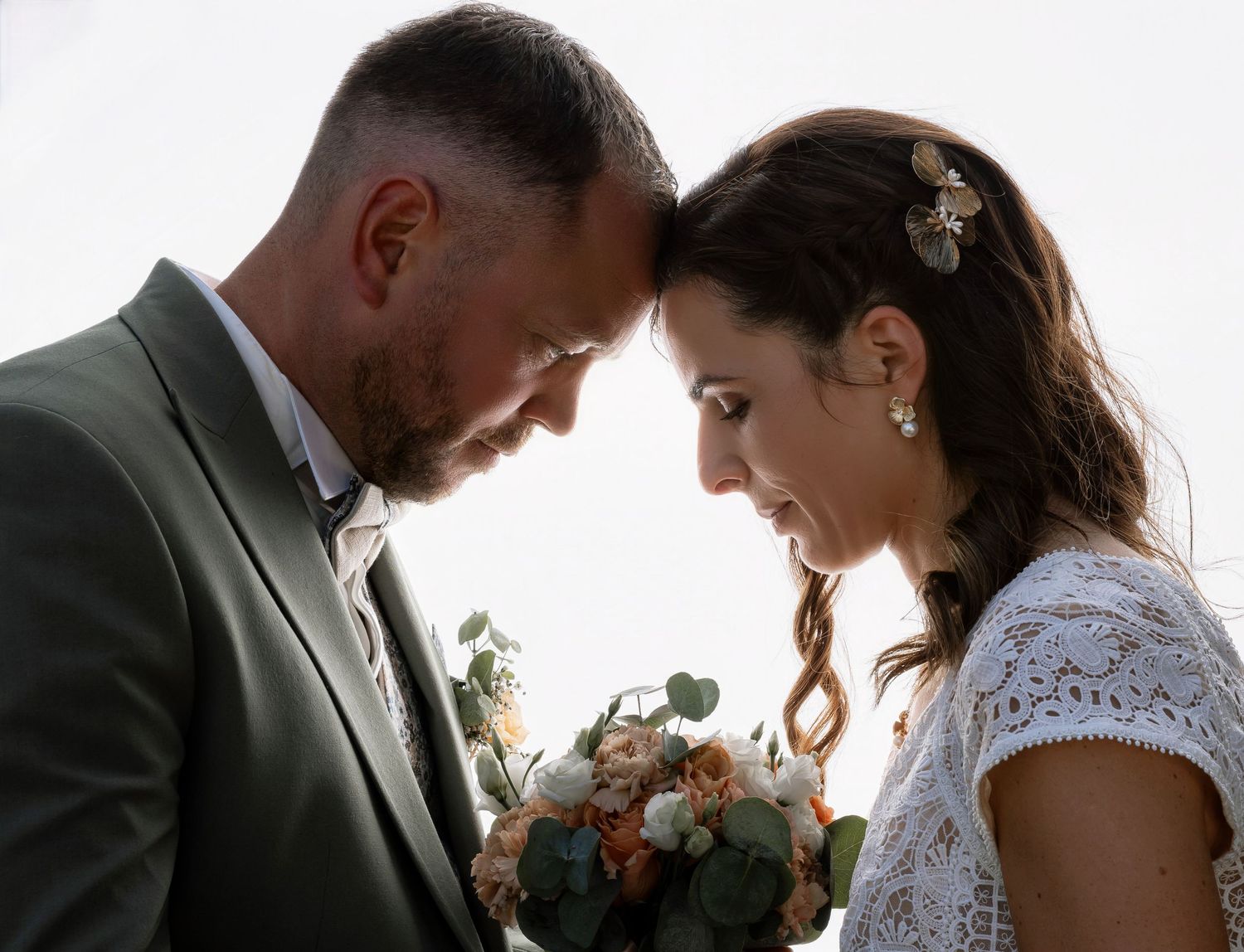 Bride and groom touching foreheads tenderly, bride holding a pastel bouquet, in an intimate outdoor wedding portrait.