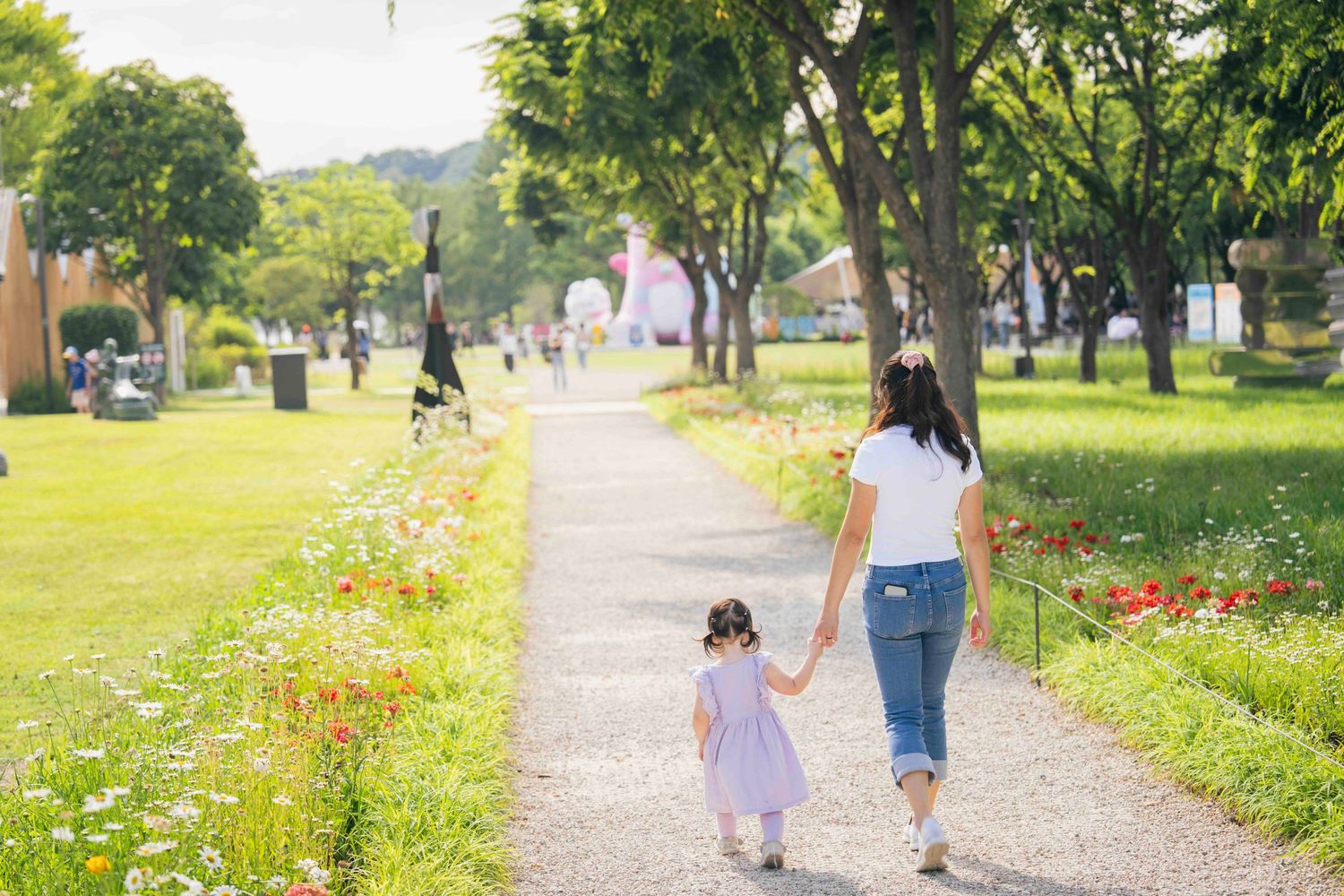 A mother and young daughter walk hand-in-hand along a flower-lined park path surrounded by lush green trees.