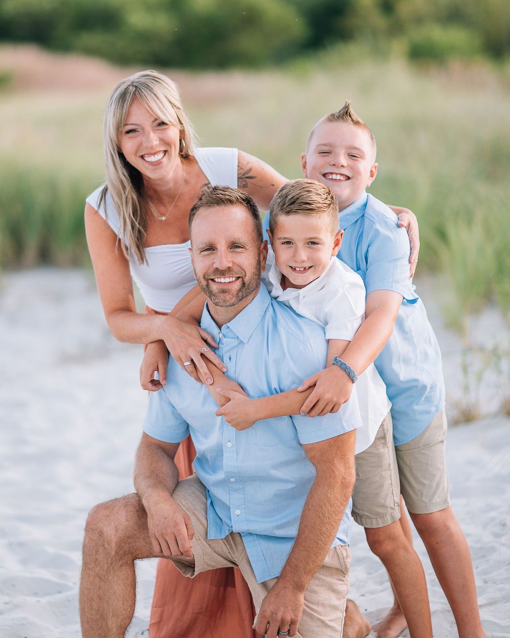 Happy family of four posing together on a sandy beach, smiling in coordinating blue and white outfits.