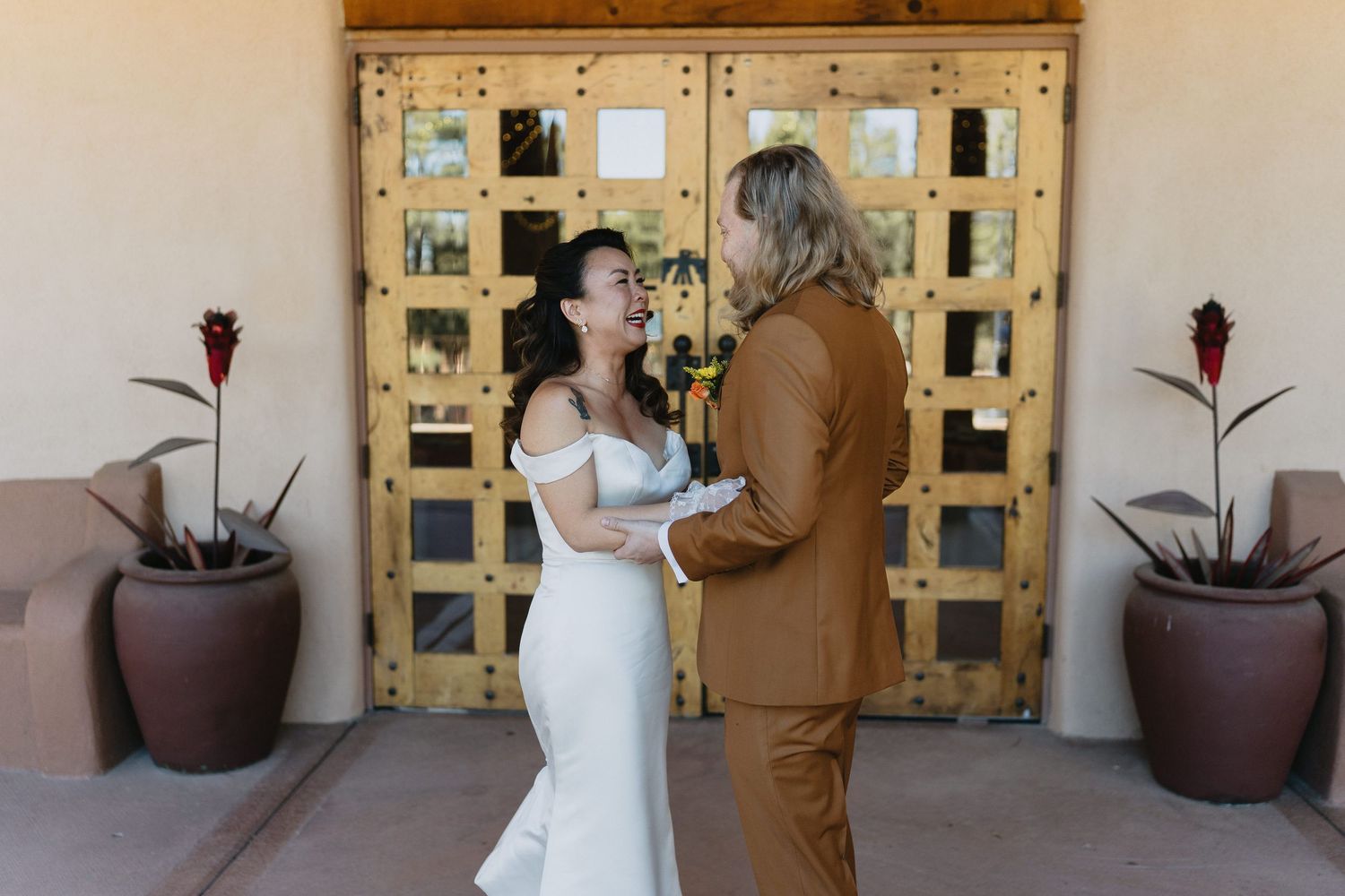 Bride in off-shoulder white gown and groom in brown suit laugh together at wedding.
