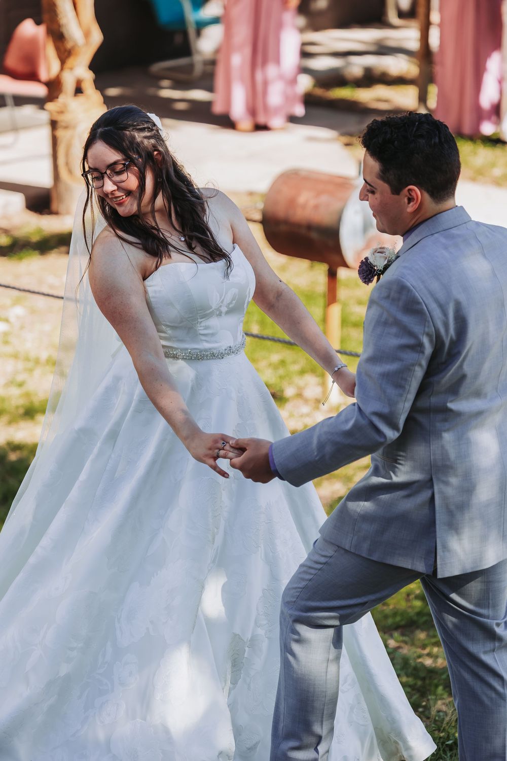 Bride in white gown and groom in gray suit hold hands and dance outdoors at a sunny wedding celebration.