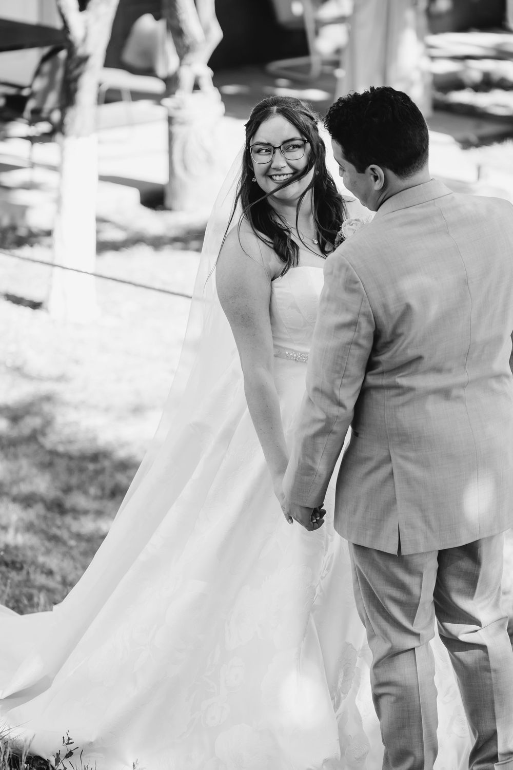 Black and white photo of a smiling bride in glasses holding hands with groom at outdoor wedding ceremony.
