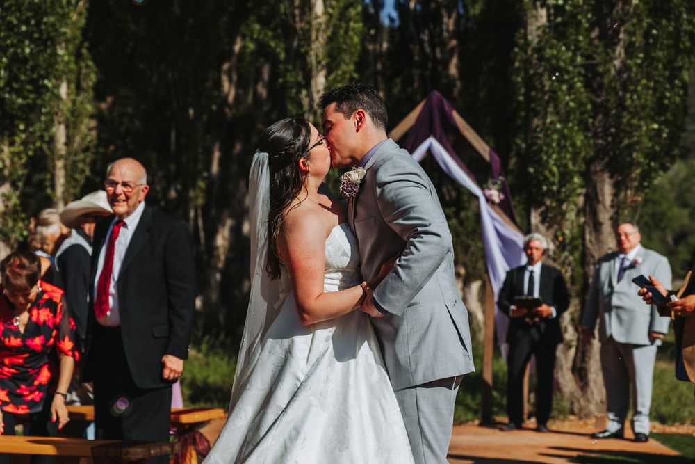 Bride and groom share their first kiss at an outdoor wedding ceremony surrounded by tall trees and guests.