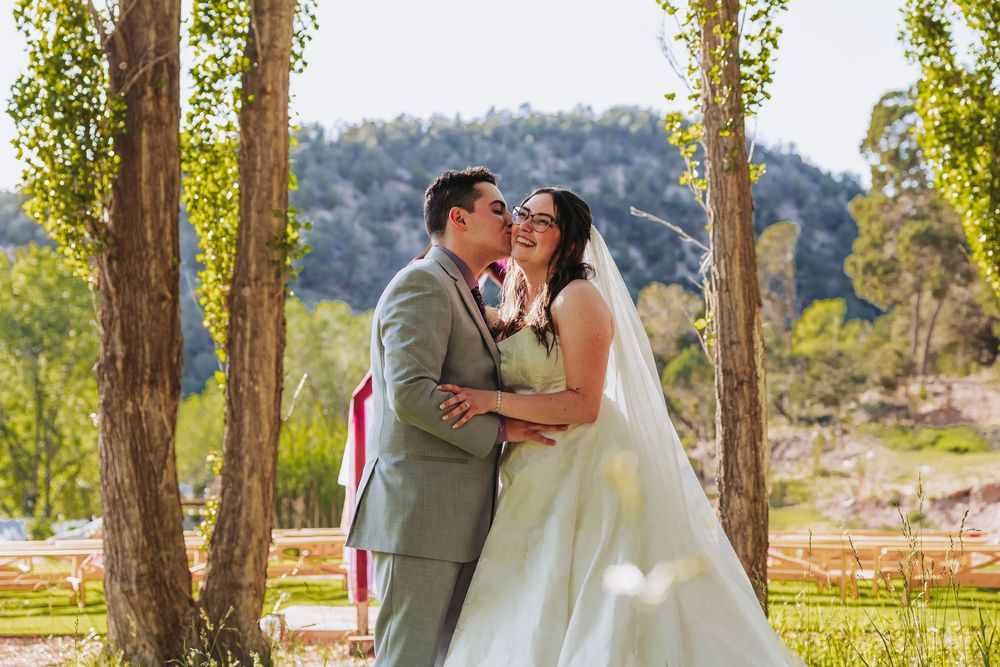 Bride and groom share a kiss under a rustic wooden arch surrounded by lush green trees at an outdoor wedding.