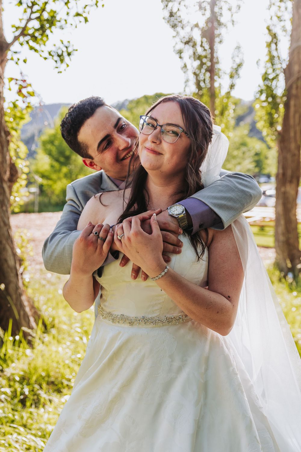 Groom in gray suit hugging bride in white gown and veil outdoors, sharing a tender moment in a lush green setting.