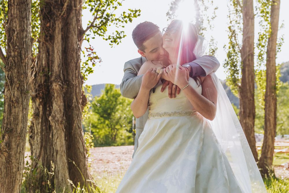 Bride and groom share a romantic embrace outdoors among trees with sunlight streaming through on their wedding day.
