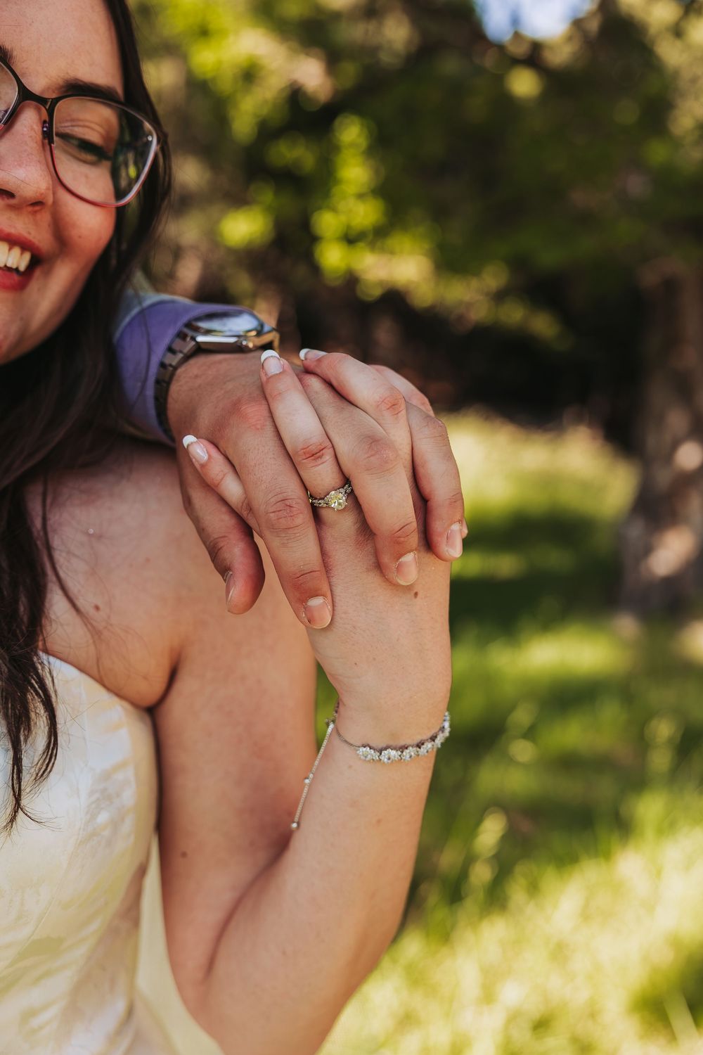 Close-up of a laughing bride and groom holding hands, showcasing wedding ring and diamond bracelet outdoors.