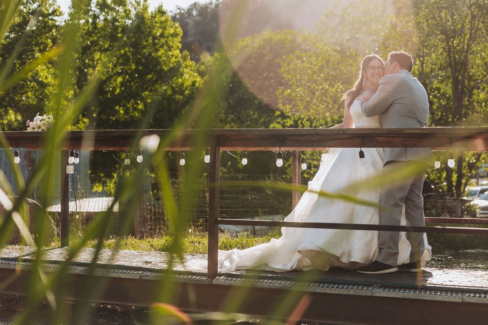 Bride and groom share a romantic embrace on a sunlit bridge surrounded by lush greenery on their wedding day.