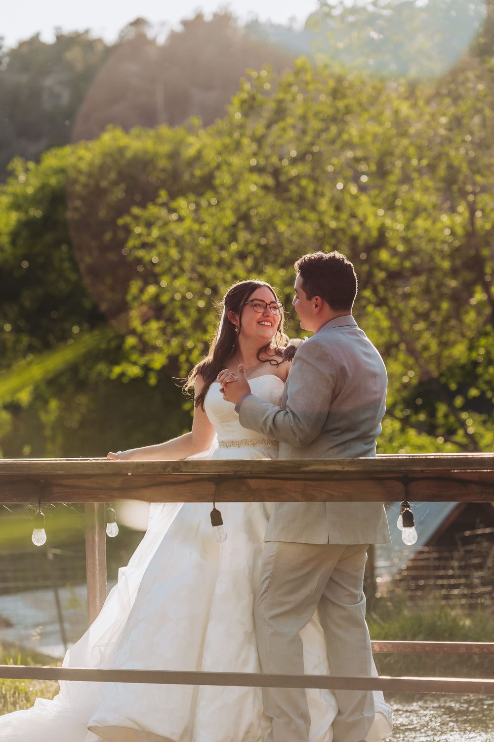 A bride and groom share a joyful moment on a sunlit wooden deck surrounded by lush greenery on their wedding day.