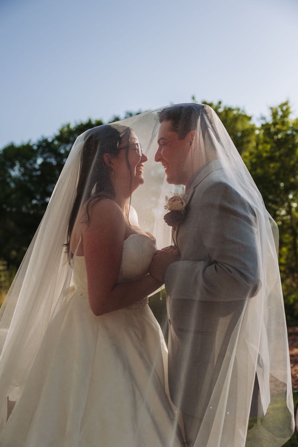 Bride and groom share a romantic moment under a flowing white veil outdoors on their wedding day.