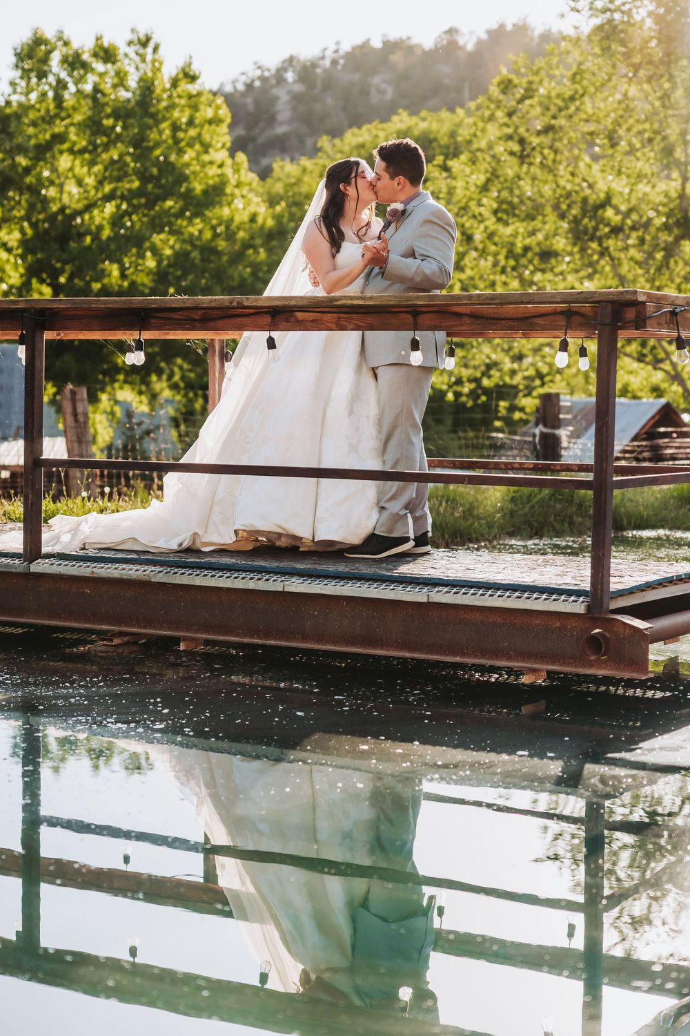 Bride and groom kissing on a rustic wooden dock over water, with their reflection visible below, surrounded by lush greenery.