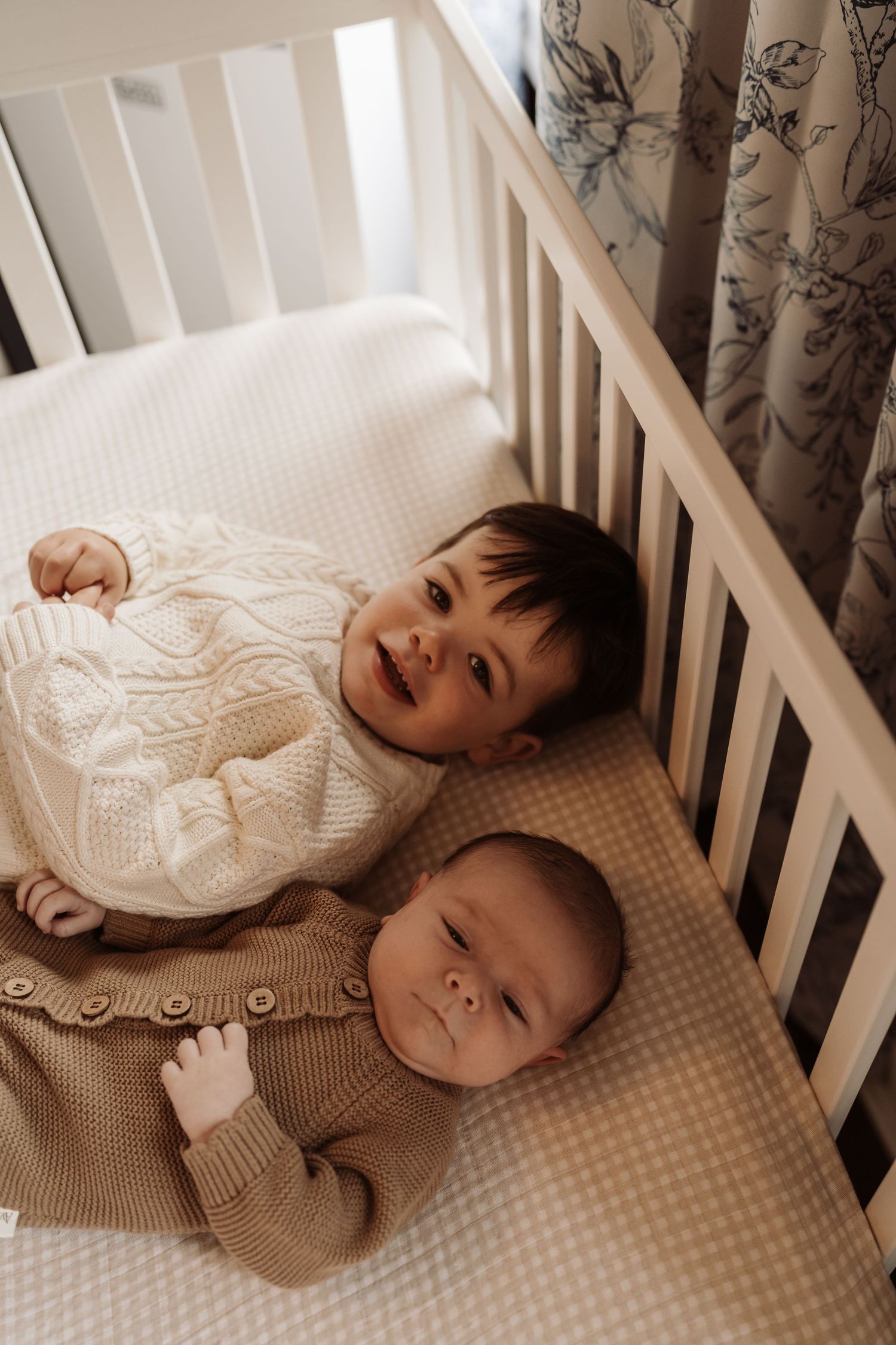 Two babies lying in a white crib, older toddler smiling in cream knit sweater beside newborn in tan cardigan.