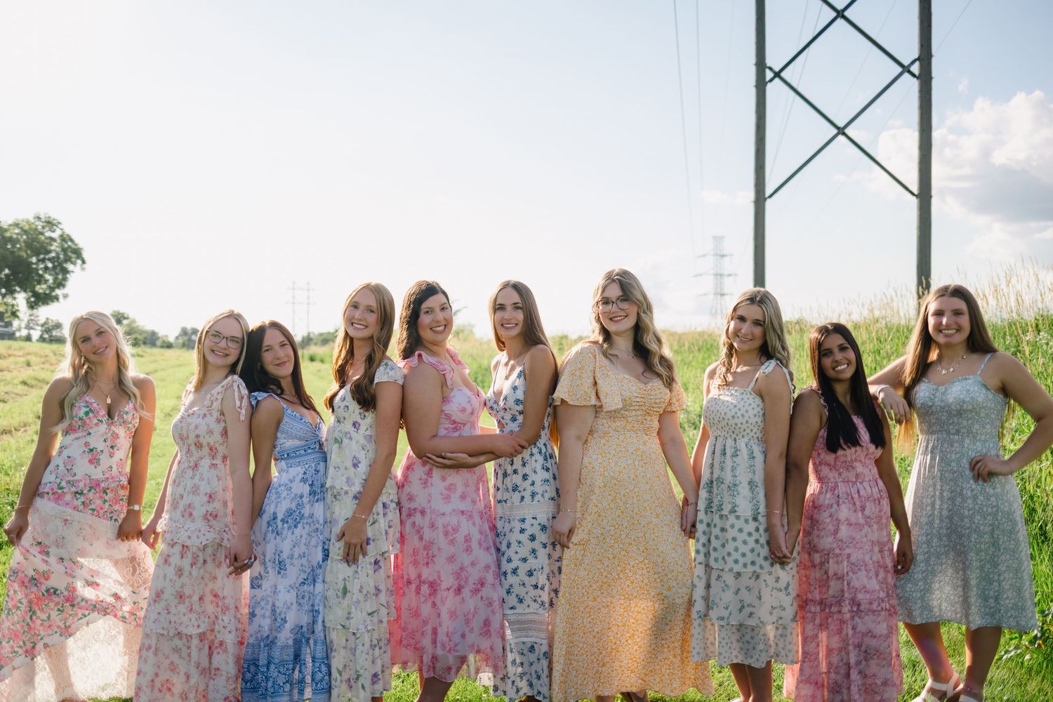 Nine women in floral dresses posing outdoors near a power tower on a sunny day.
