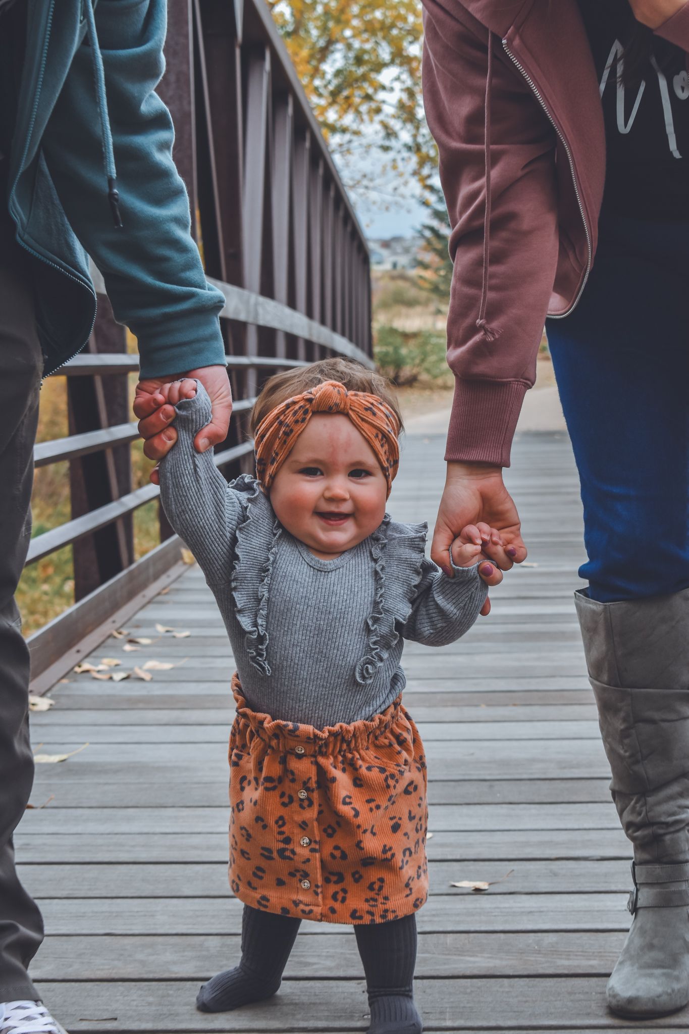 Smiling baby girl in orange leopard skirt and headband holds parents hands while standing on a wooden bridge in autumn.