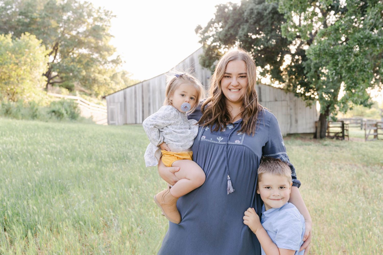 A smiling woman in a gray dress holds a toddler while a young boy stands beside her in a sunny green field.
