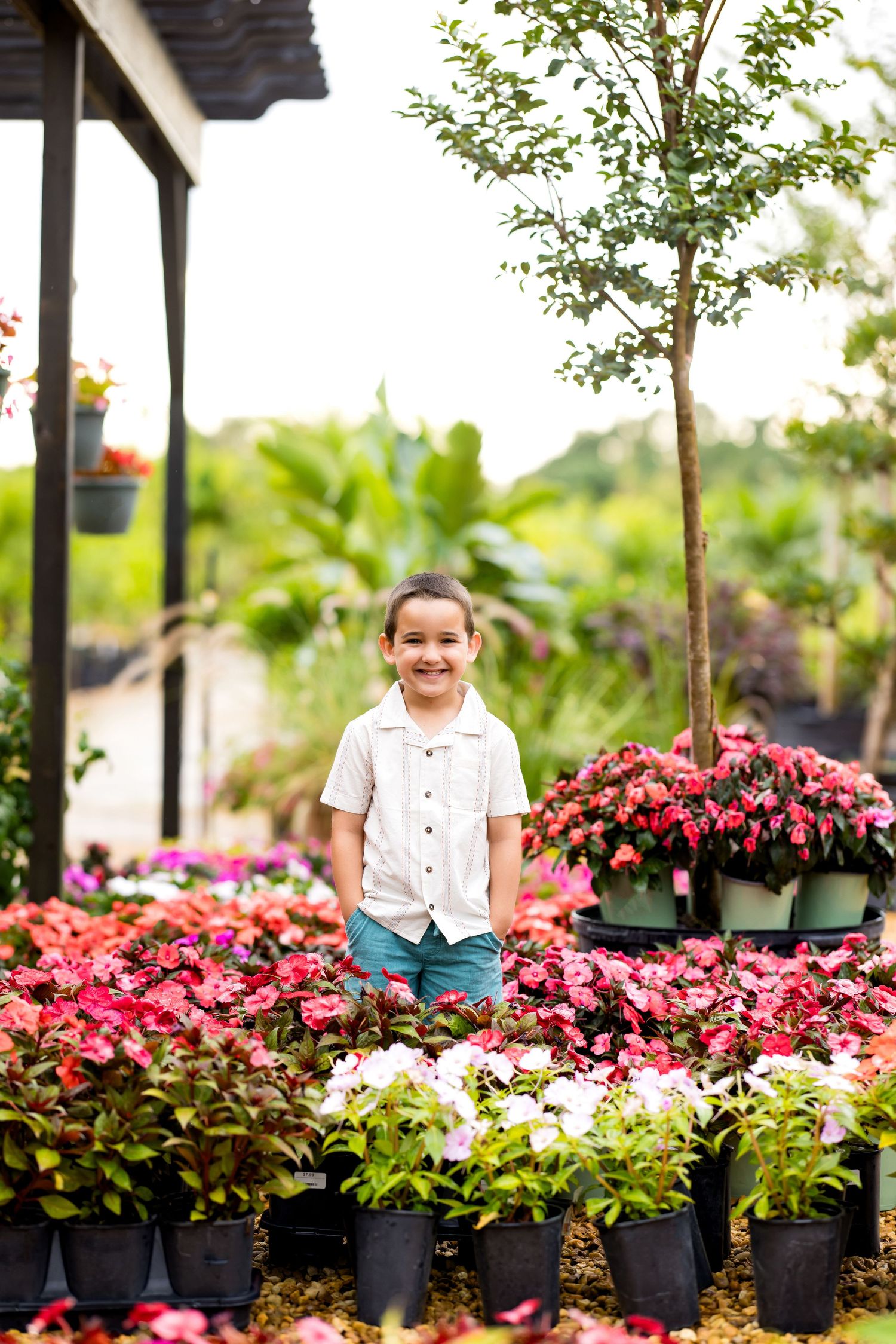 A smiling child in a white shirt stands among colorful potted flowers at an outdoor garden nursery.