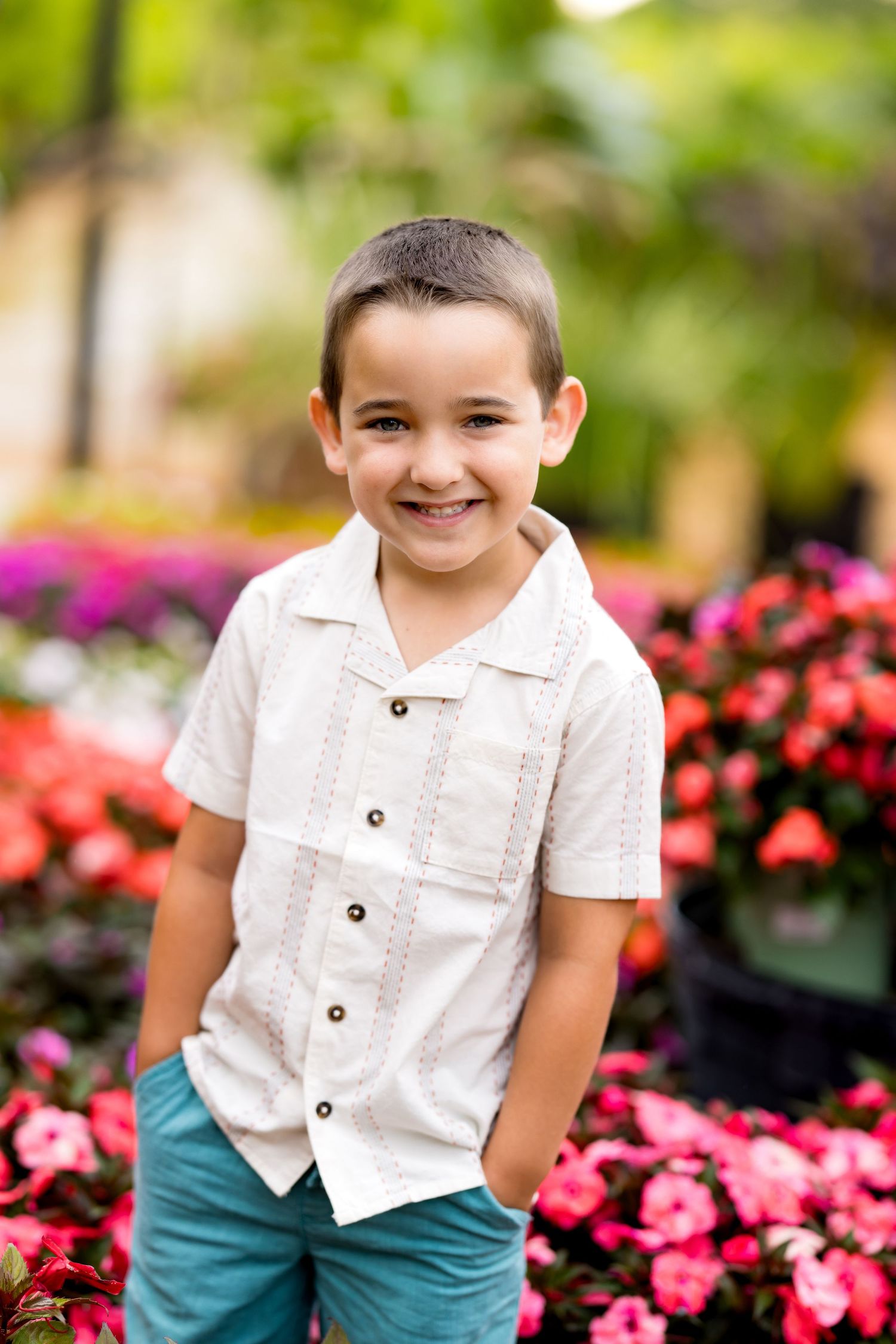 Smiling young boy in white button-up shirt and teal pants standing in front of colorful garden flowers.
