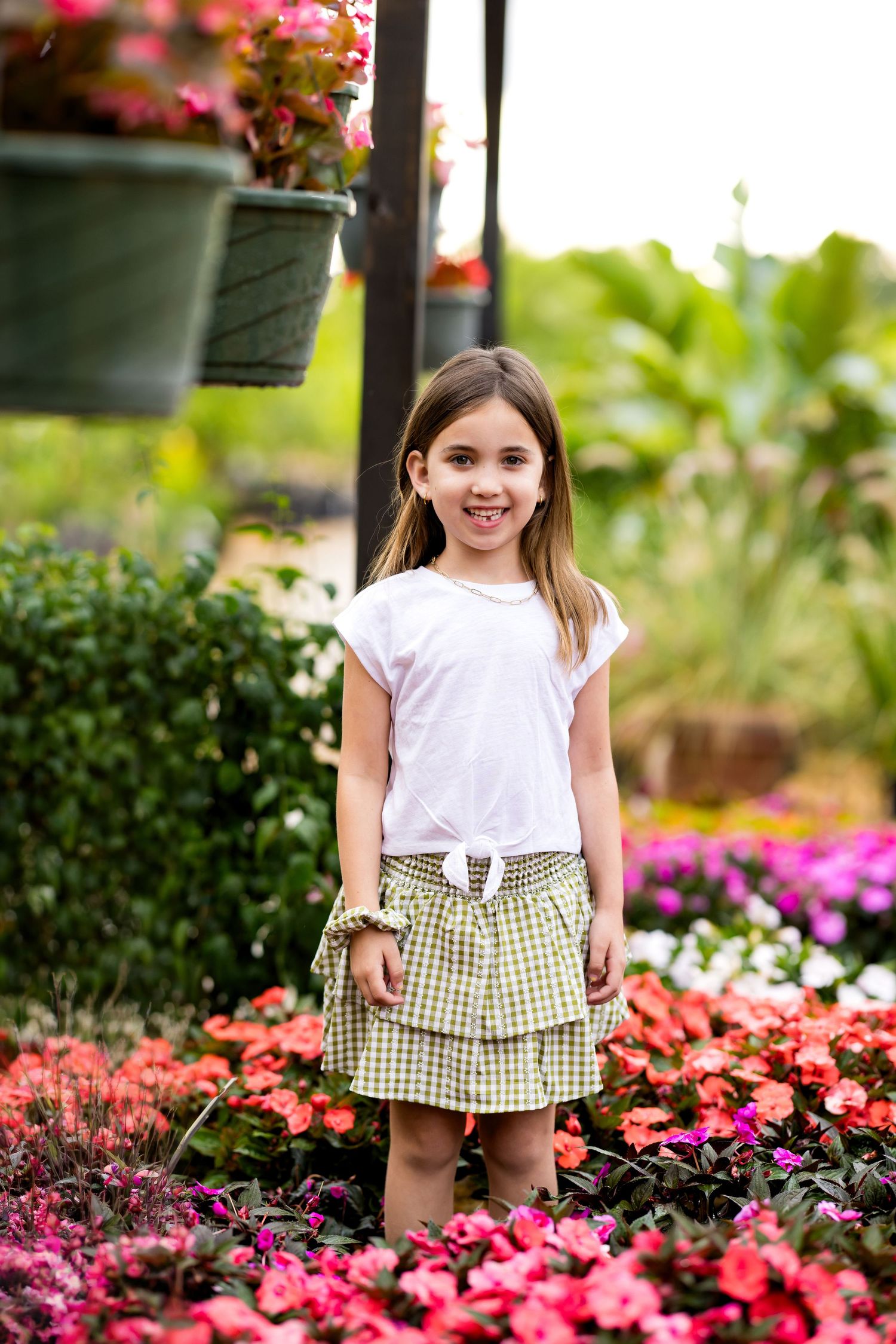 A young girl in a white top and plaid skirt smiles among colorful flowers in a garden nursery.