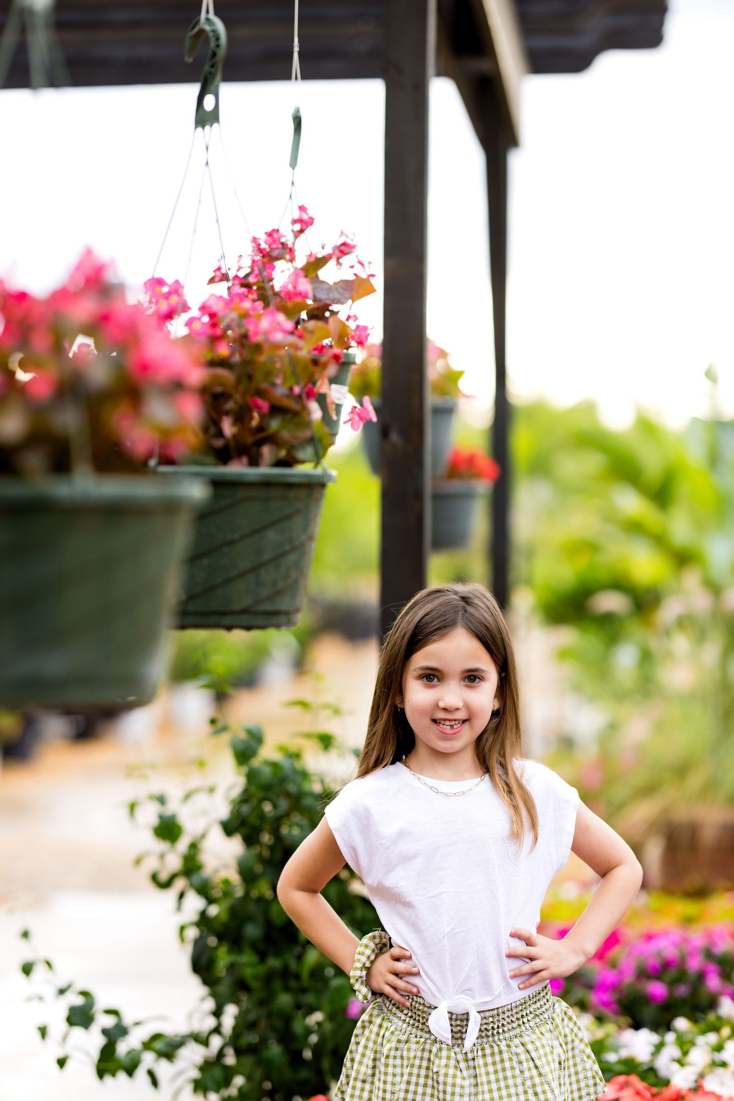 Young girl in white top and plaid skirt poses confidently in a colorful garden nursery with hanging flower baskets.