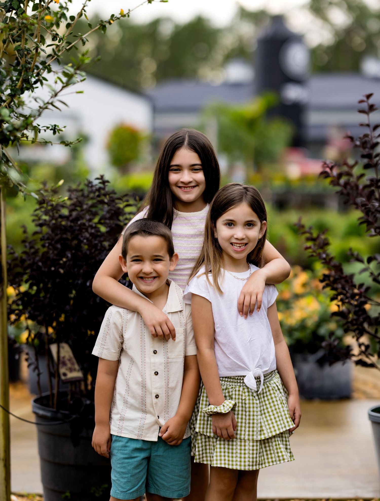 Three smiling children pose together outdoors among lush green plants and trees at a garden nursery.