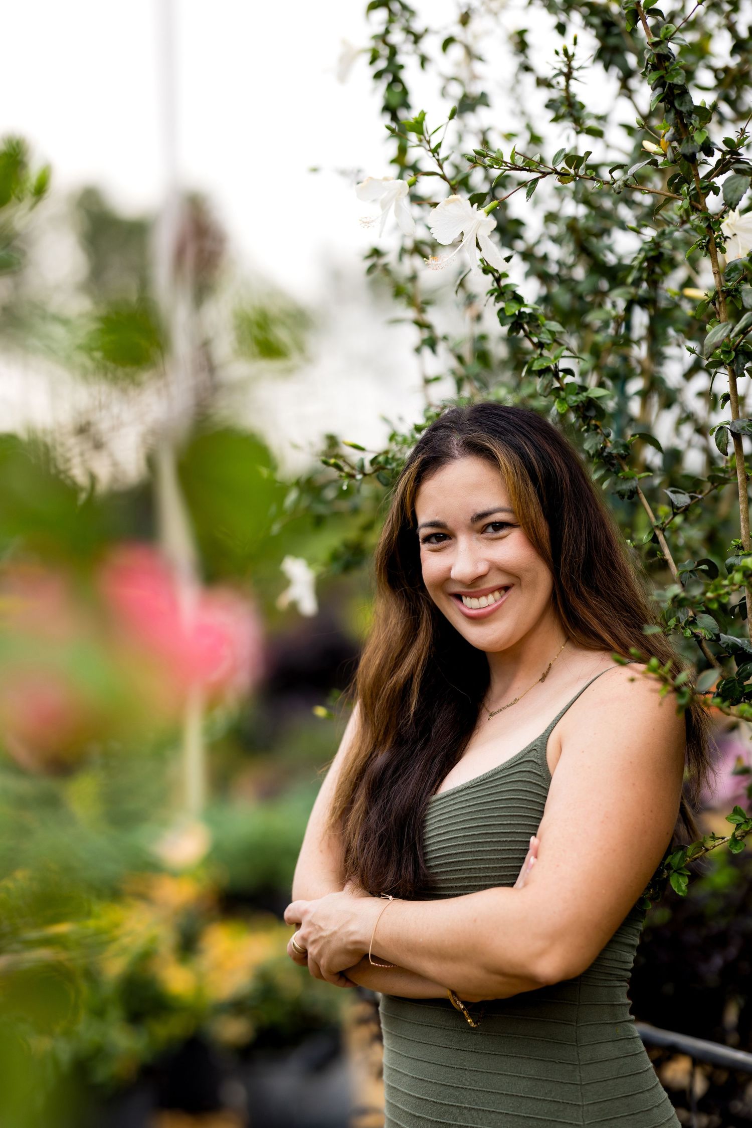 A smiling woman in a green dress stands with crossed arms among vibrant plants in a garden.