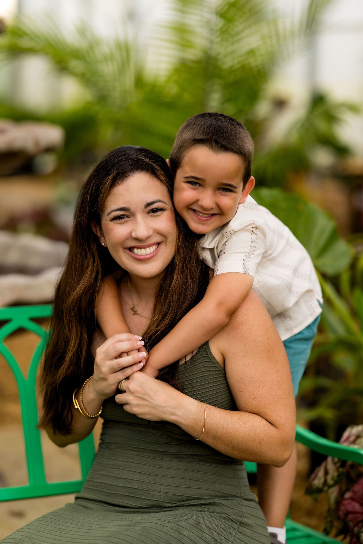 A smiling woman in a green dress hugs a young boy in a greenhouse setting.