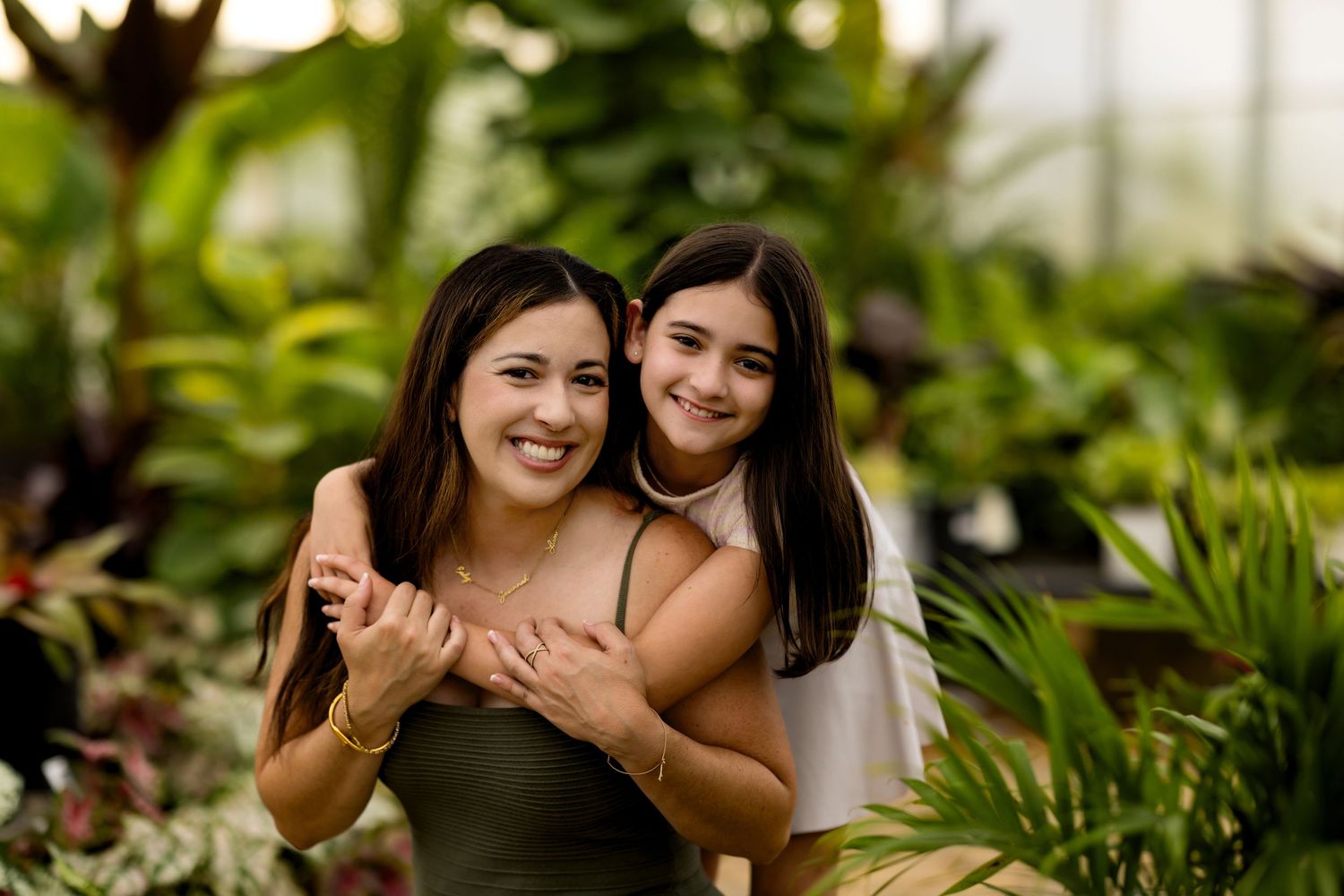 Two women smiling and hugging in a lush green greenhouse, surrounded by tropical plants.