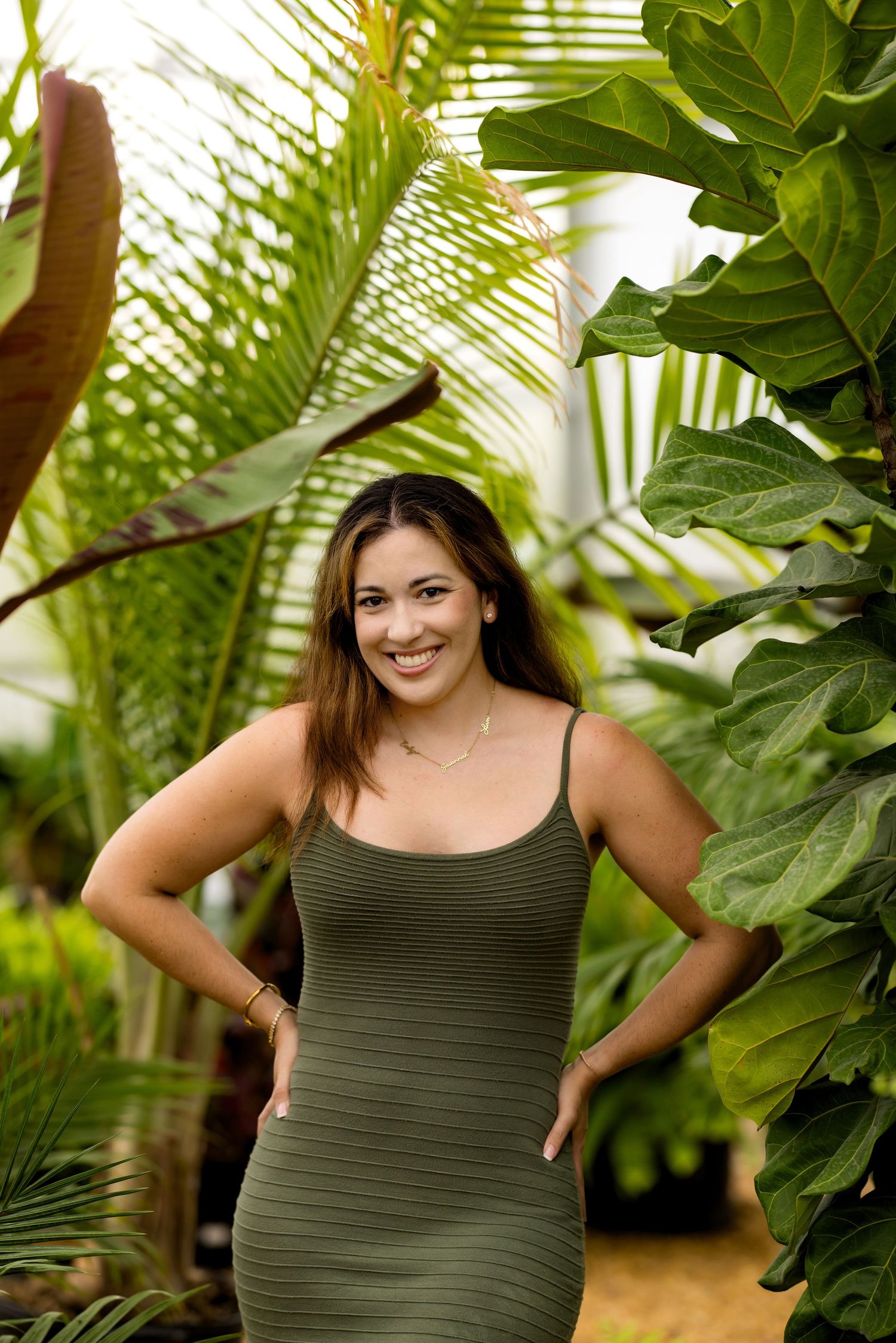 Woman in olive green ribbed dress smiling among tropical plants in a lush greenhouse setting.