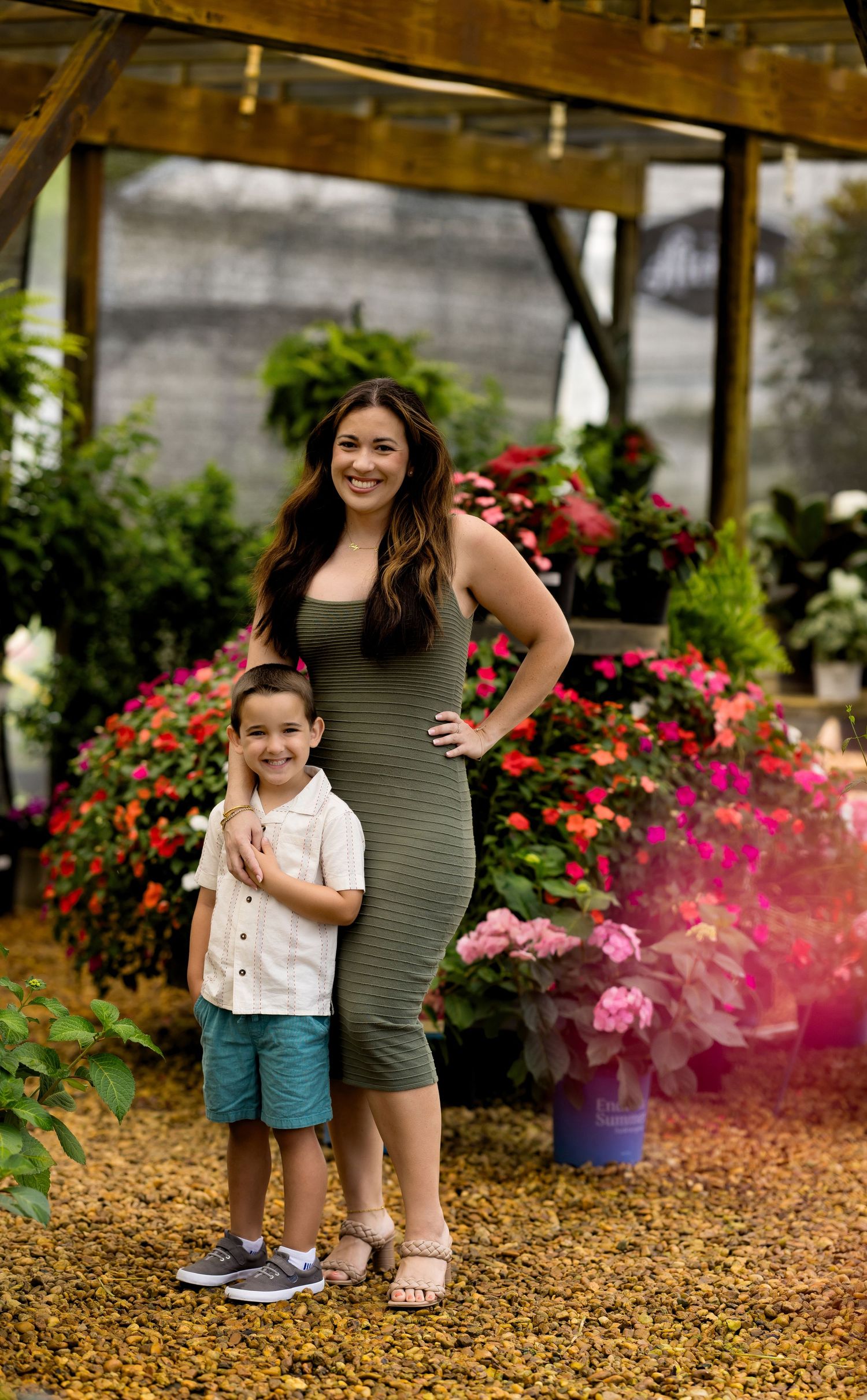 A woman in an olive dress and young boy in white shirt pose together at a colorful flower garden nursery.