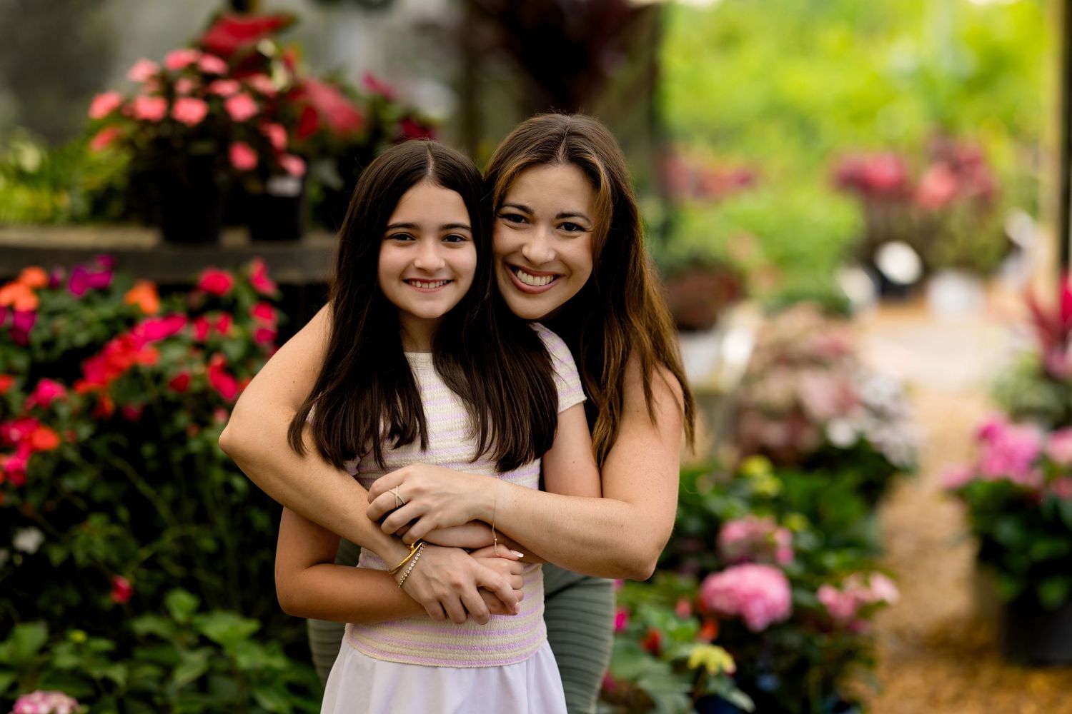 Two smiling girls embrace in a colorful flower garden, wearing white dresses among vibrant pink and red blooms.