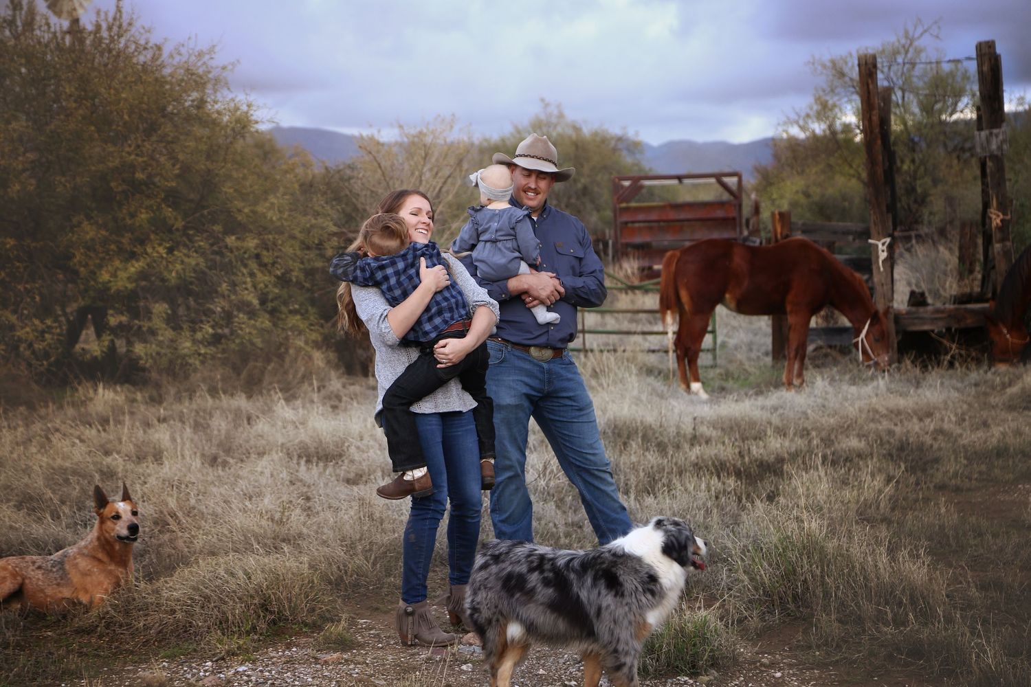 Family with two children, a cowboy hat, two dogs and a brown horse standing on a rustic ranch at dusk.