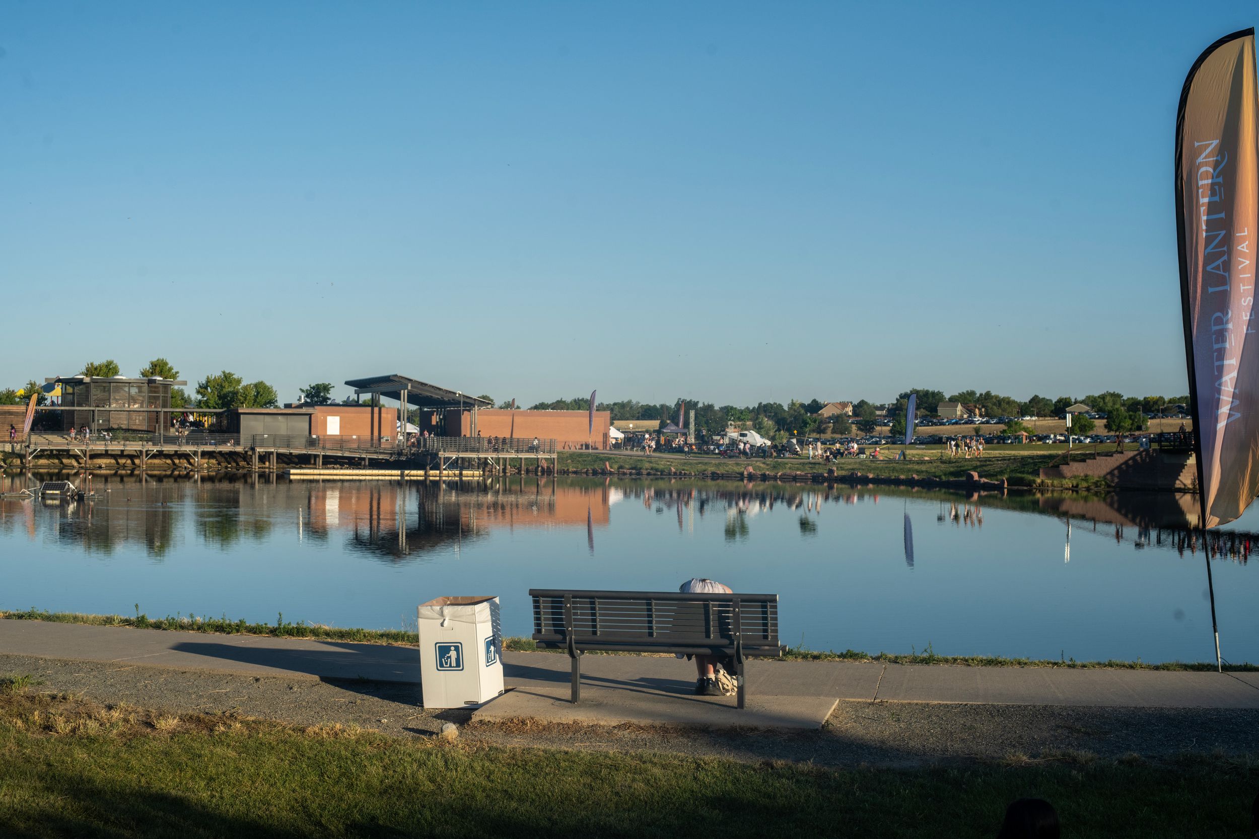 A serene harbor waterfront at dusk with a bench, reflective calm water, buildings, and decorative flags under a clear blue sky.
