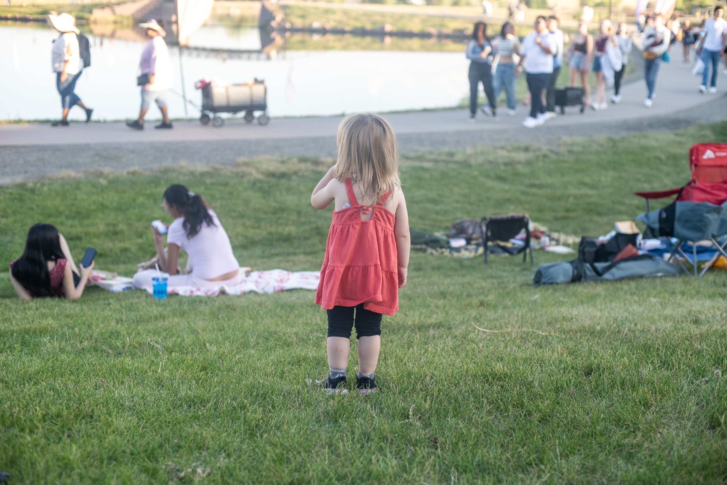 Young girl in red dress standing on green grass at an outdoor public event near a waterfront.