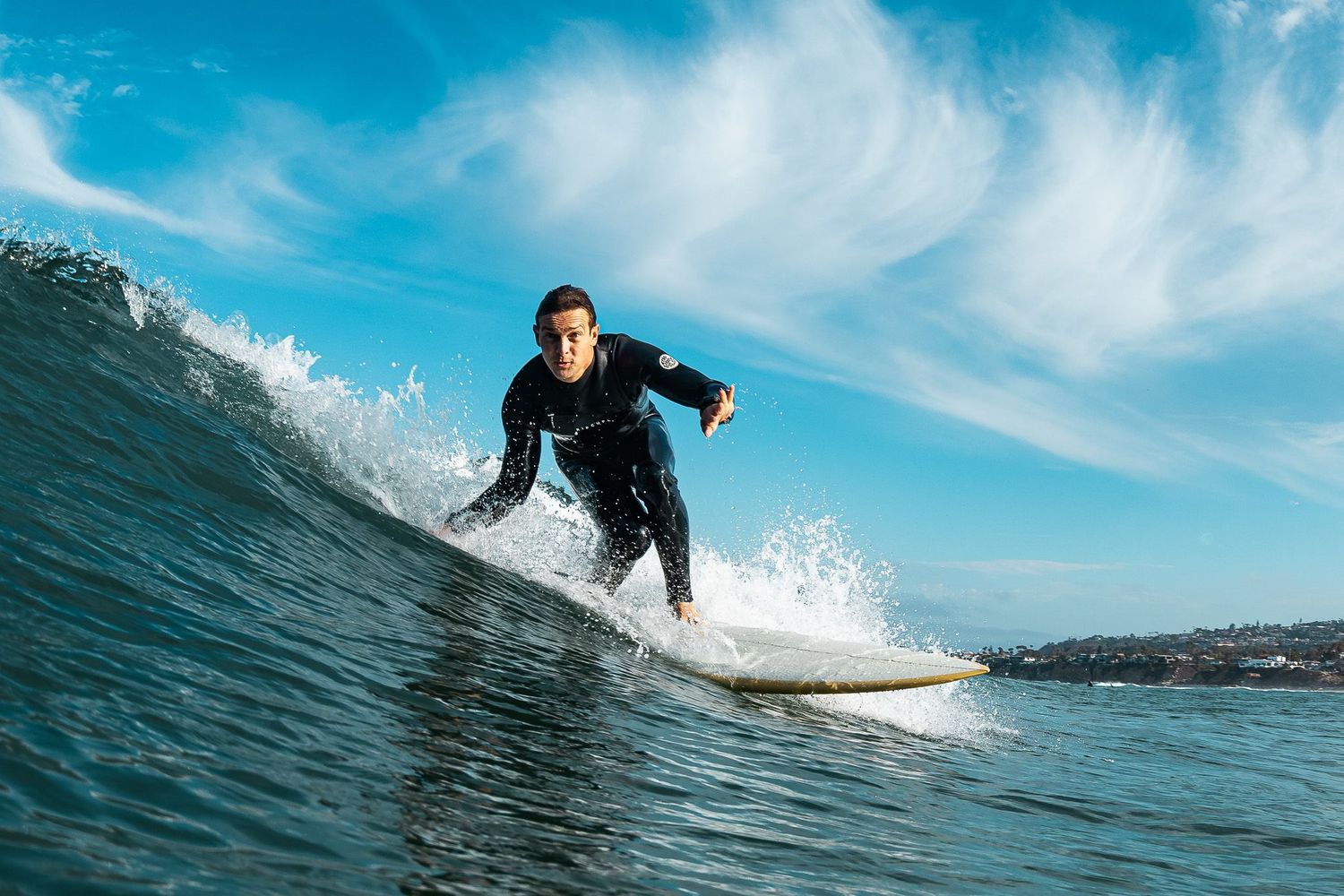 San Diego surf photography of a male surfer riding an ocean wave in a black wetsuit against a blue sky with wispy clouds.