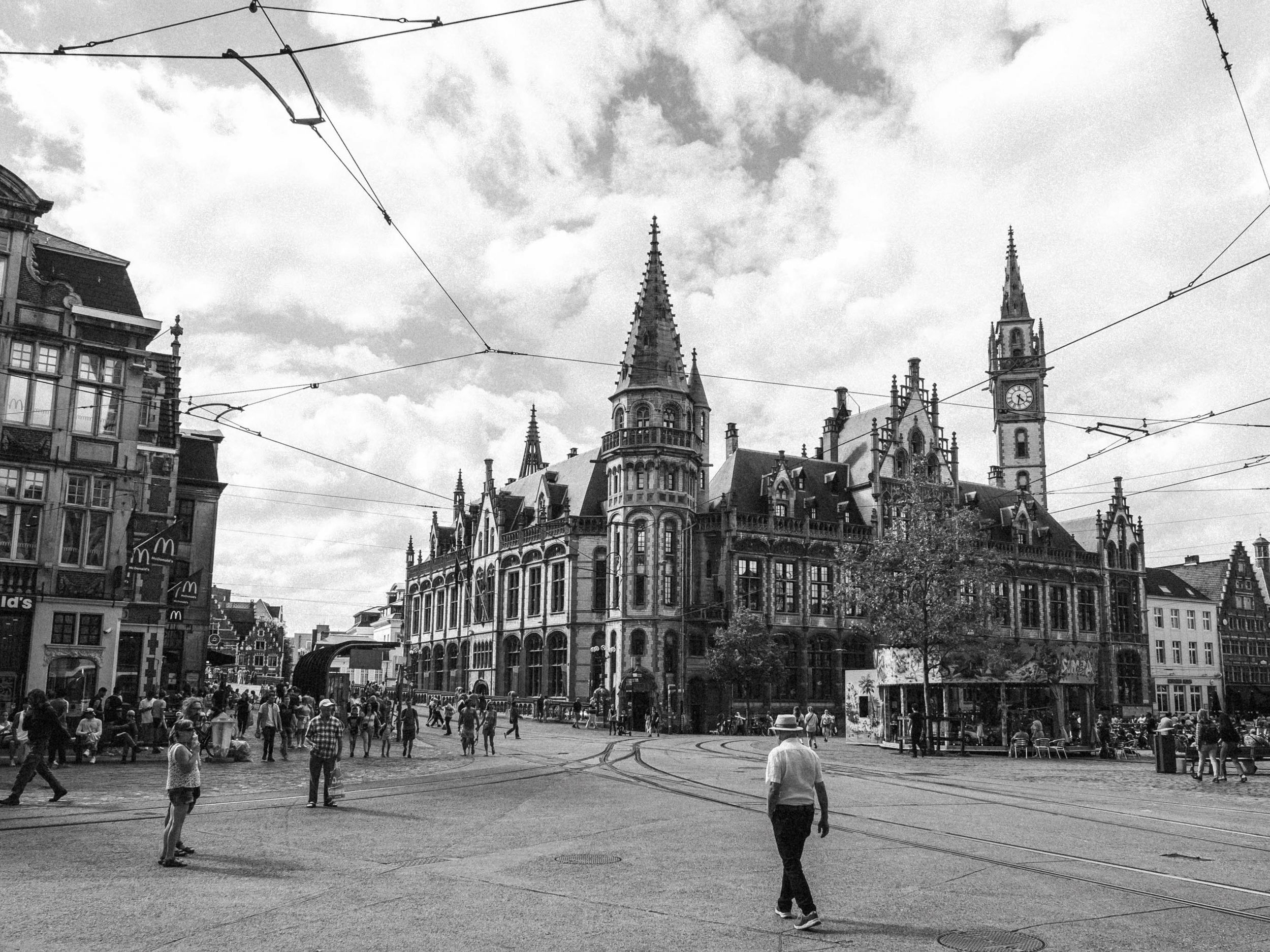 Černobílá městská scéna s historickou architekturou, tramvajovými kolejemi a chodci.
