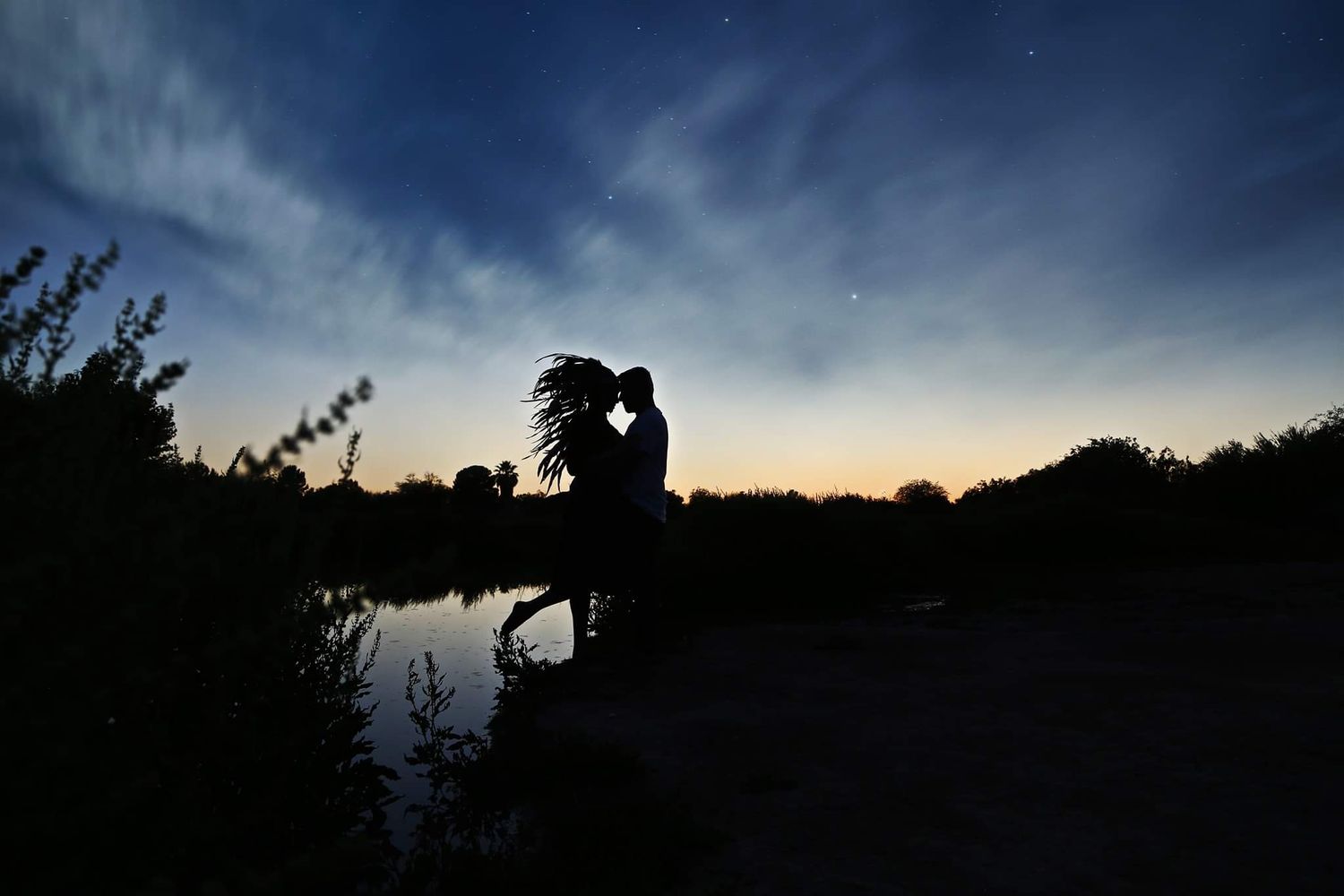 Silhouette of a couple embracing beside a calm lake under a deep blue twilight sky with soft clouds and a glowing horizon.