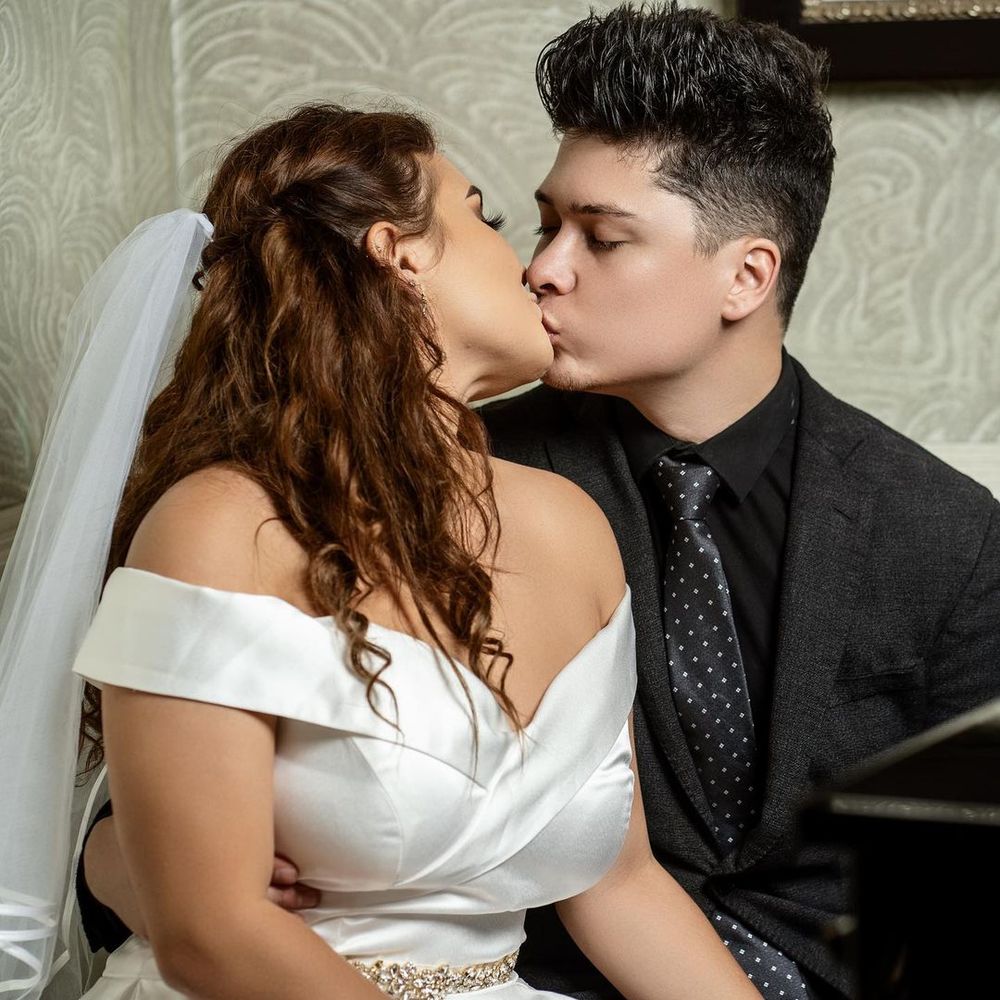 A bride in a white off-shoulder gown and veil shares a romantic kiss with a groom in a dark suit and tie.