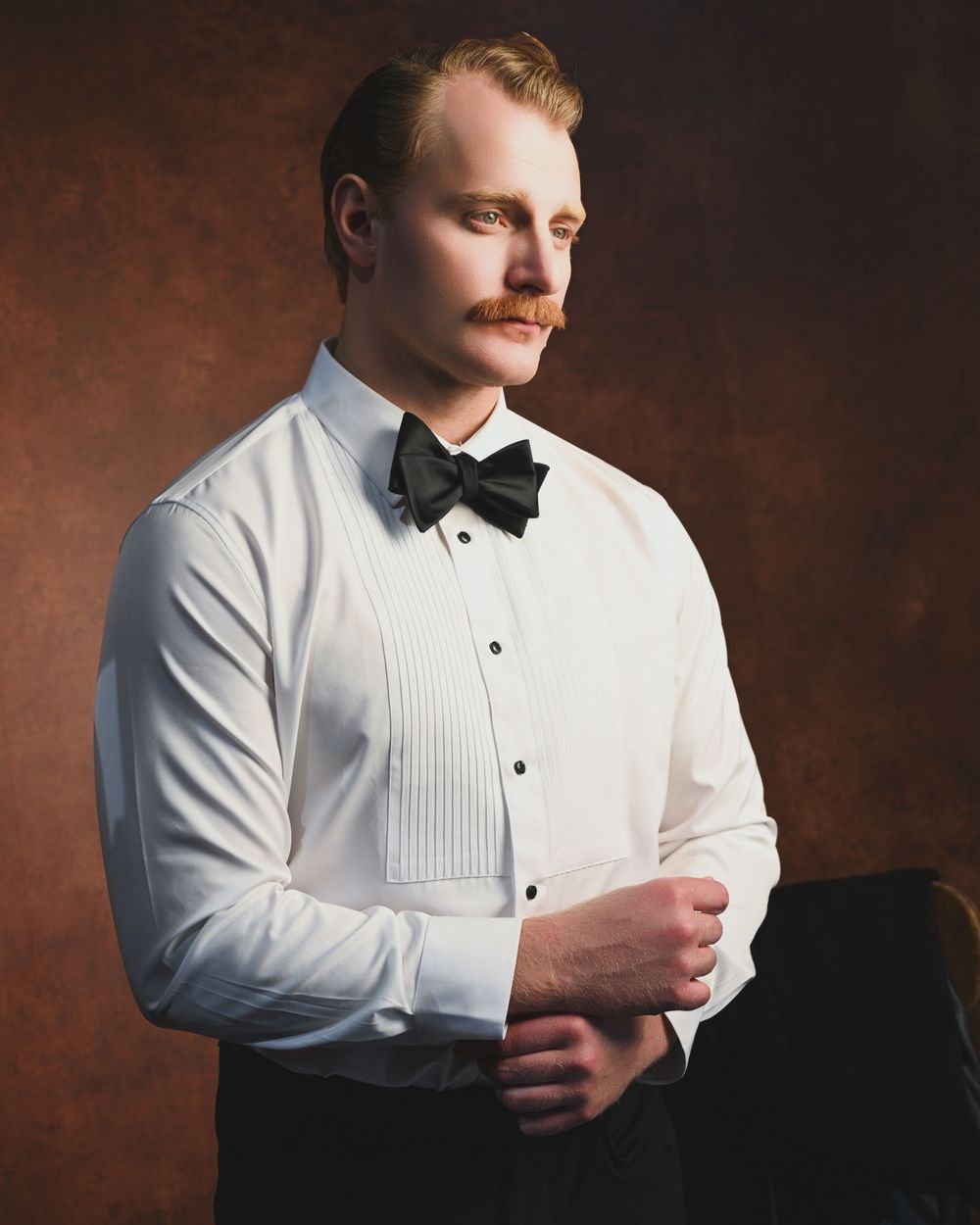 Man in white tuxedo shirt and black bow tie adjusting cufflinks, styled portrait with vintage aesthetic.