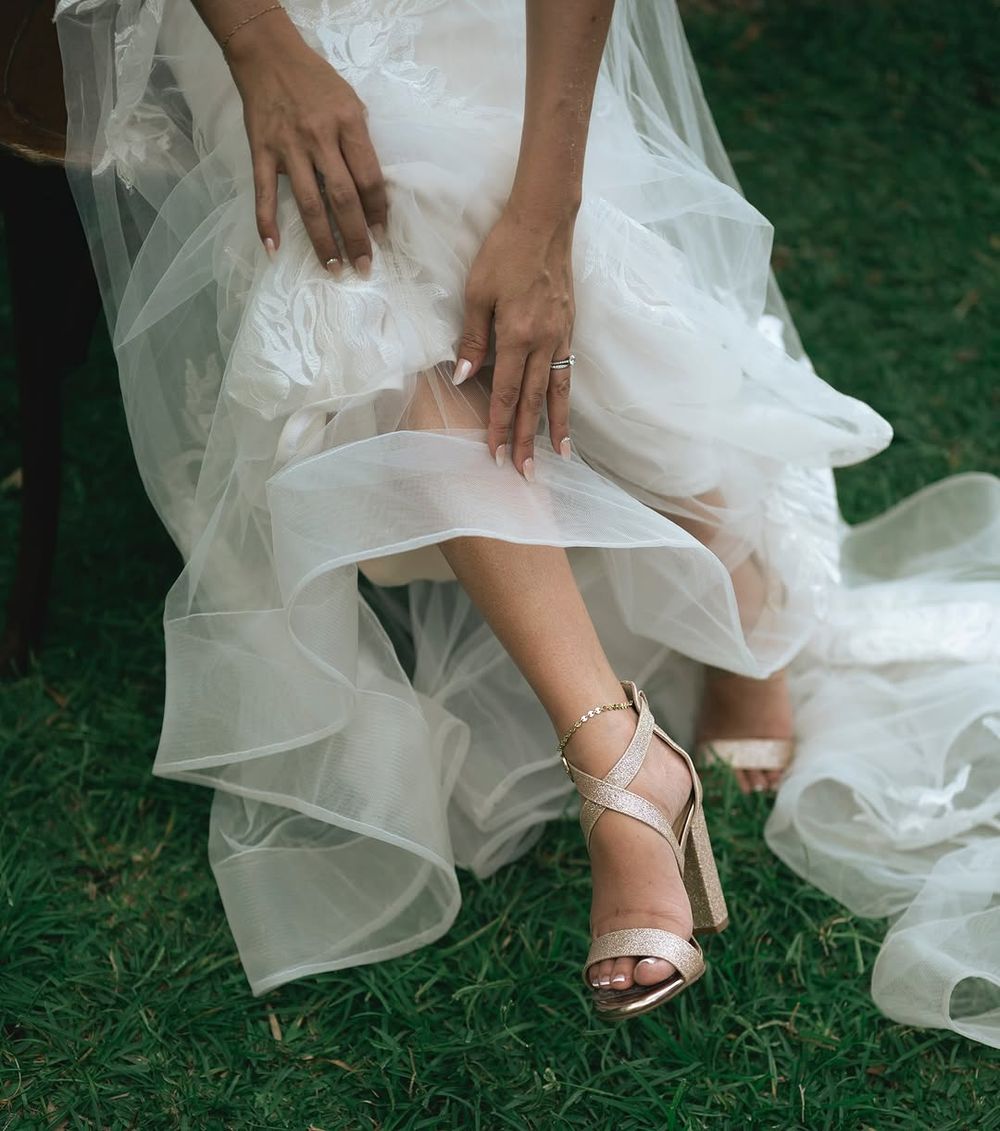 Bride in white tulle gown adjusting rose gold ankle strap heels while seated on green grass.