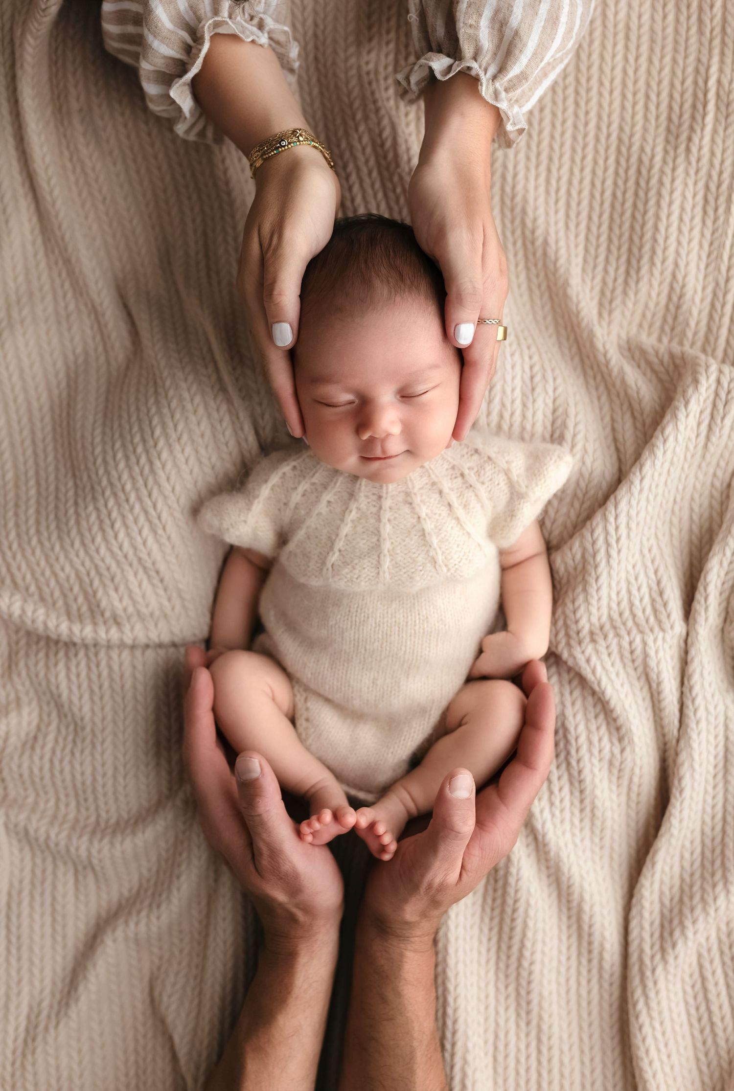 Newborn girl dressed in a soft romper smiles gently as mom and dad cradle her with their hands, captured by Charlottesville photographer.