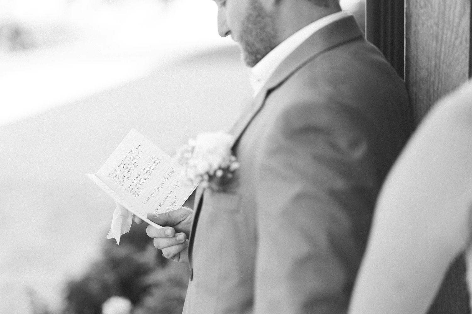 Groom in suit reads wedding vows from paper, wearing boutonniere, black and white photo.