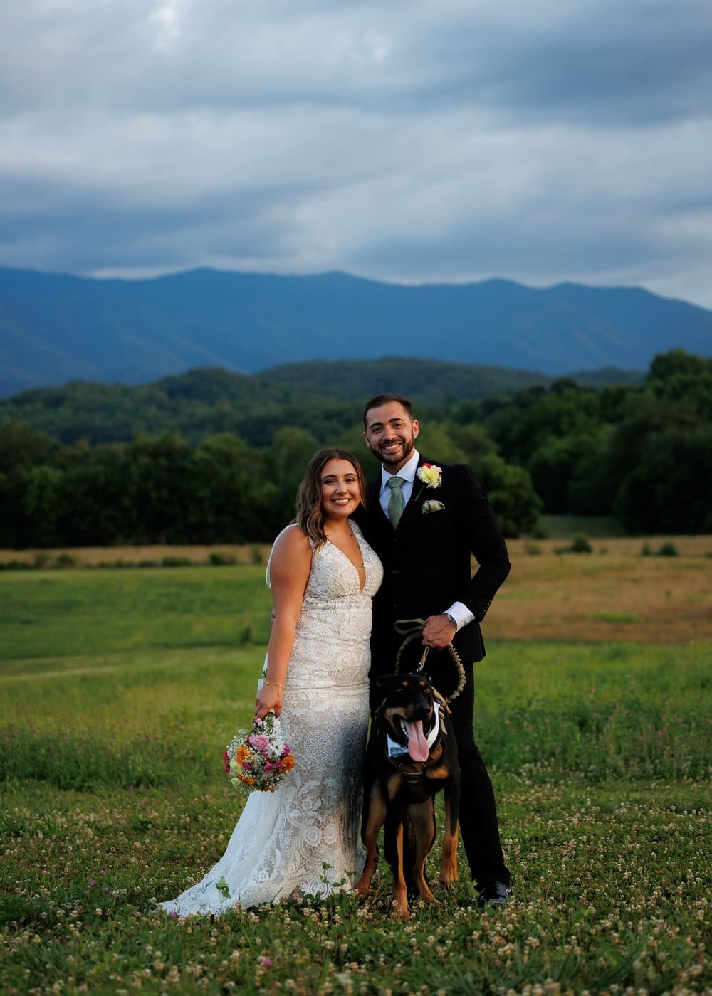 Bride and groom pose with their German Shepherd dog in a green meadow with mountain views on their wedding day.