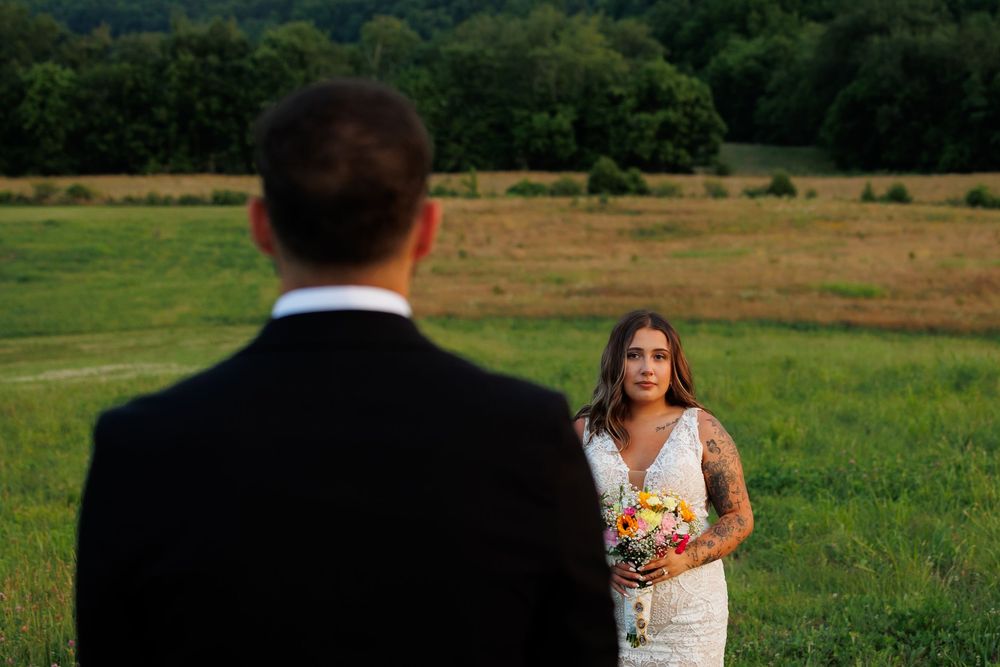 Bride in lace gown holding colorful bouquet stands in green field facing groom in black tuxedo during outdoor wedding.