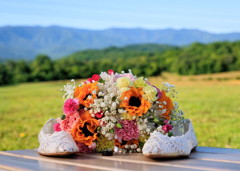 Colorful wedding bouquet with orange, pink, and white flowers beside lace bridal shoes on a table with mountain backdrop.