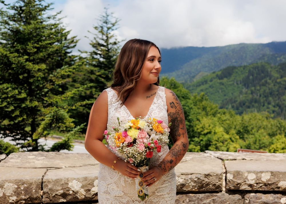 Bride in lace wedding dress holding colorful bouquet with mountain forest backdrop.
