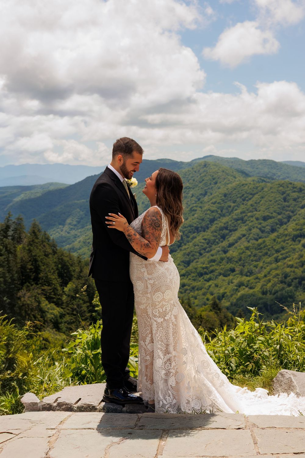Bride and groom share romantic moment on stone overlook surrounded by lush green mountains.