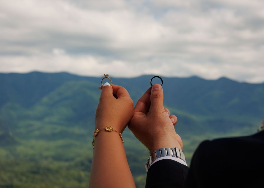 Couple holding wedding rings up against a scenic mountain landscape with lush green hills and cloudy sky.